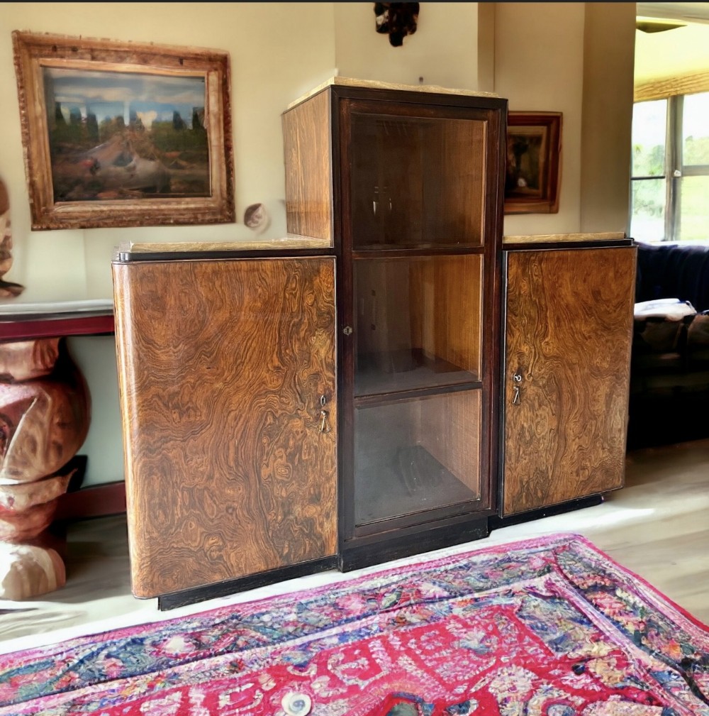 An Italian Art Deco tiered cabinet featuring a central glazed compartment, twin side cupboards with figured burr walnut veneer doors, and marble tops.