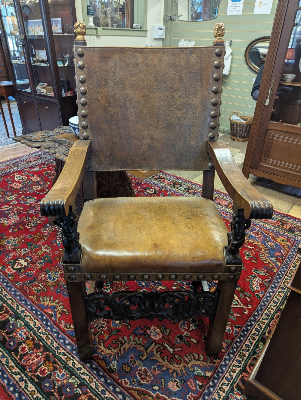 An 18th-century European walnut throne chair featuring high-backed studded leather upholstery, carved armrests, finials, and an ornate lower stretcher.