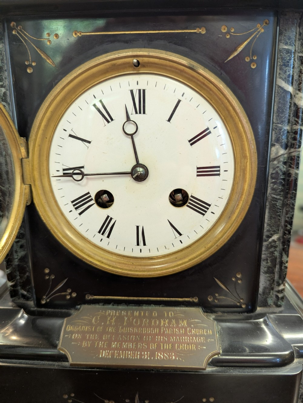 A Victorian black marble and slate mantel clock featuring incised gilded floral patterns, a white enamel dial with Roman numerals, and a brass presentation plaque. View IV