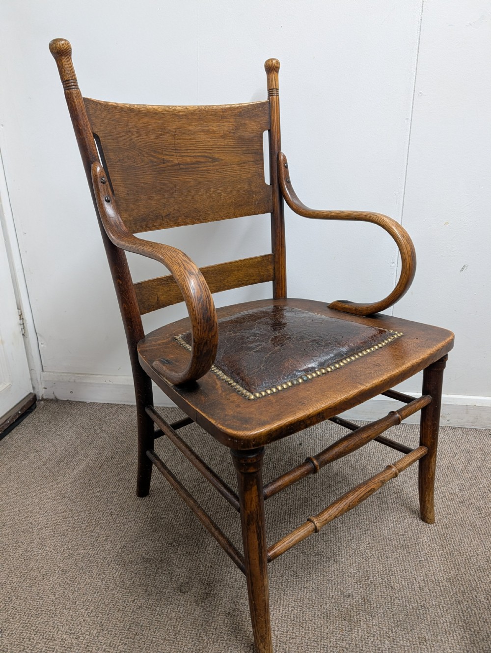 An Arts and Crafts oak open armchair featuring curved sweeping arms, a shaped timber backboard, turned legs with double stretchers, and an inset brown leather seat fixed with brass studs.