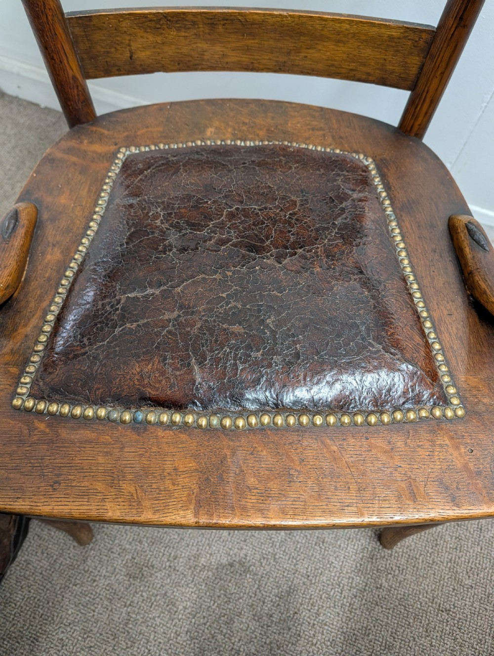 An Arts and Crafts oak open armchair featuring curved sweeping arms, a shaped timber backboard, turned legs with double stretchers, and an inset brown leather seat fixed with brass studs. View II