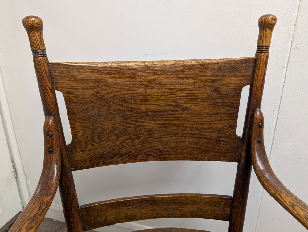 An Arts and Crafts oak open armchair featuring curved sweeping arms, a shaped timber backboard, turned legs with double stretchers, and an inset brown leather seat fixed with brass studs. View III