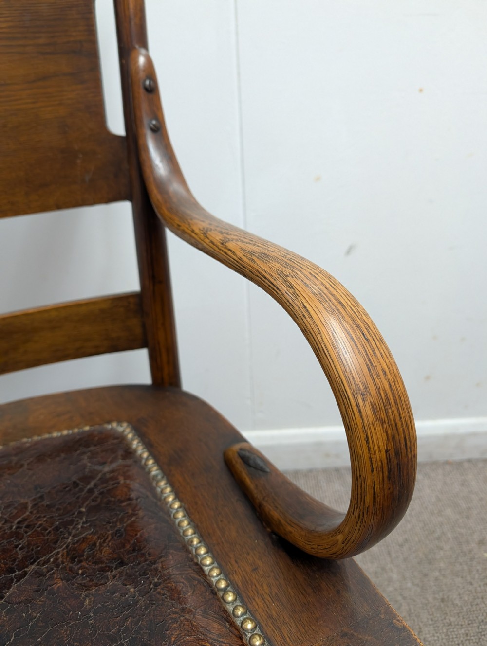 An Arts and Crafts oak open armchair featuring curved sweeping arms, a shaped timber backboard, turned legs with double stretchers, and an inset brown leather seat fixed with brass studs. View VI