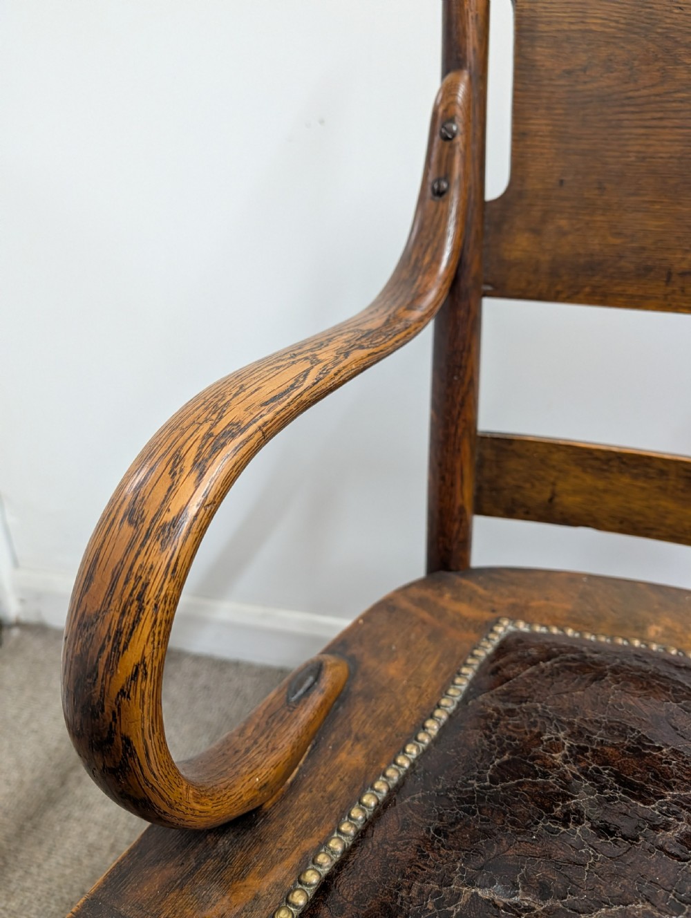 An Arts and Crafts oak open armchair featuring curved sweeping arms, a shaped timber backboard, turned legs with double stretchers, and an inset brown leather seat fixed with brass studs. View VII