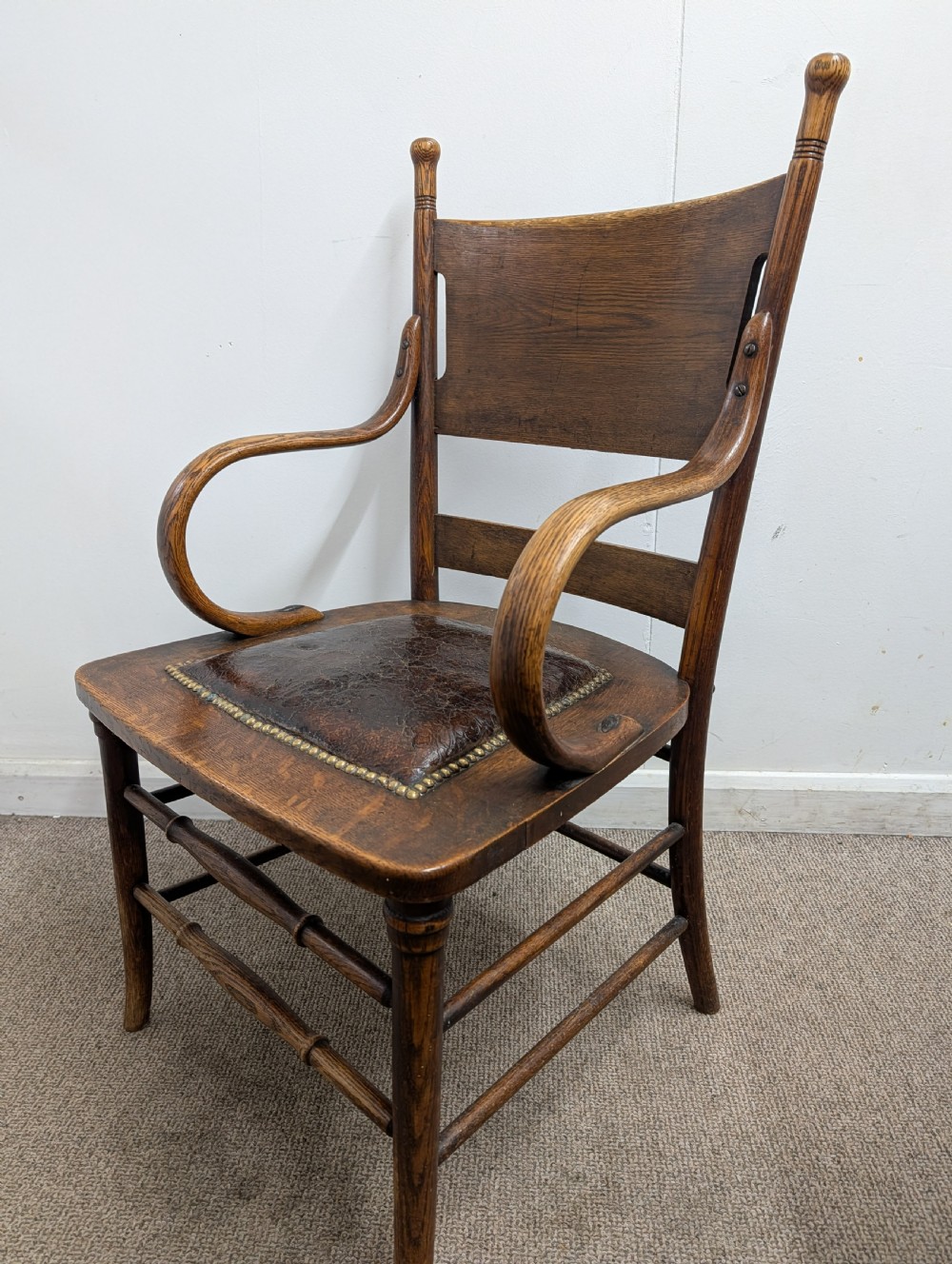 An Arts and Crafts oak open armchair featuring curved sweeping arms, a shaped timber backboard, turned legs with double stretchers, and an inset brown leather seat fixed with brass studs. View IX