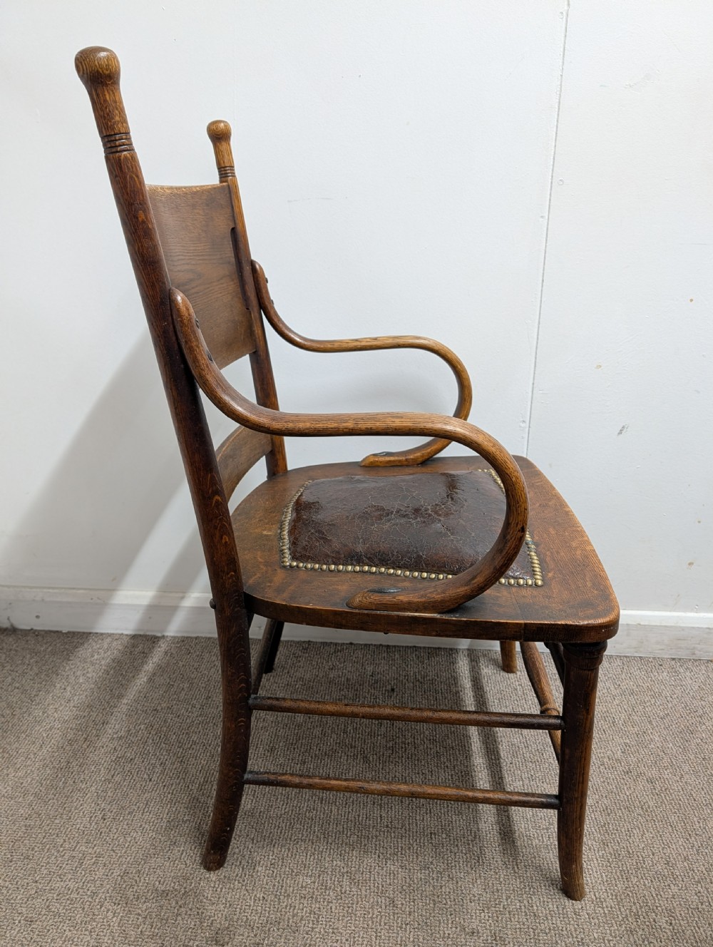 An Arts and Crafts oak open armchair featuring curved sweeping arms, a shaped timber backboard, turned legs with double stretchers, and an inset brown leather seat fixed with brass studs. View X