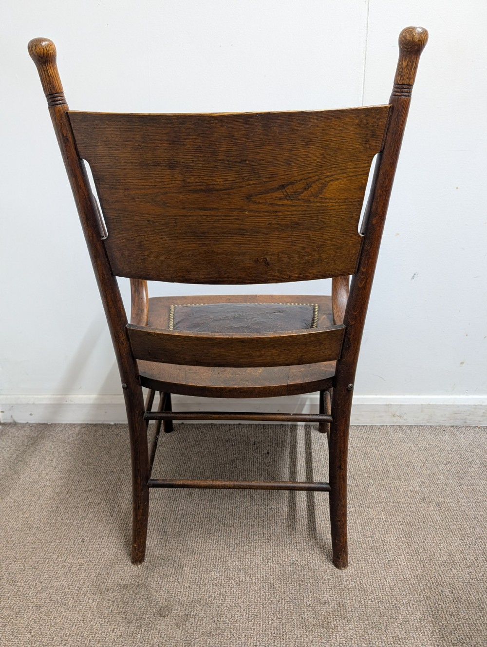 An Arts and Crafts oak open armchair featuring curved sweeping arms, a shaped timber backboard, turned legs with double stretchers, and an inset brown leather seat fixed with brass studs. View XI