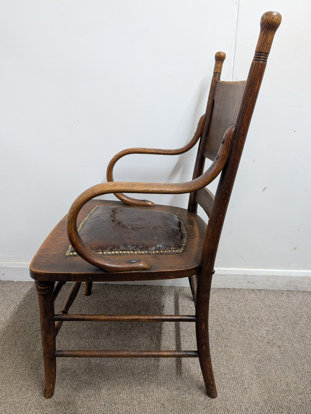 An Arts and Crafts oak open armchair featuring curved sweeping arms, a shaped timber backboard, turned legs with double stretchers, and an inset brown leather seat fixed with brass studs. View XII