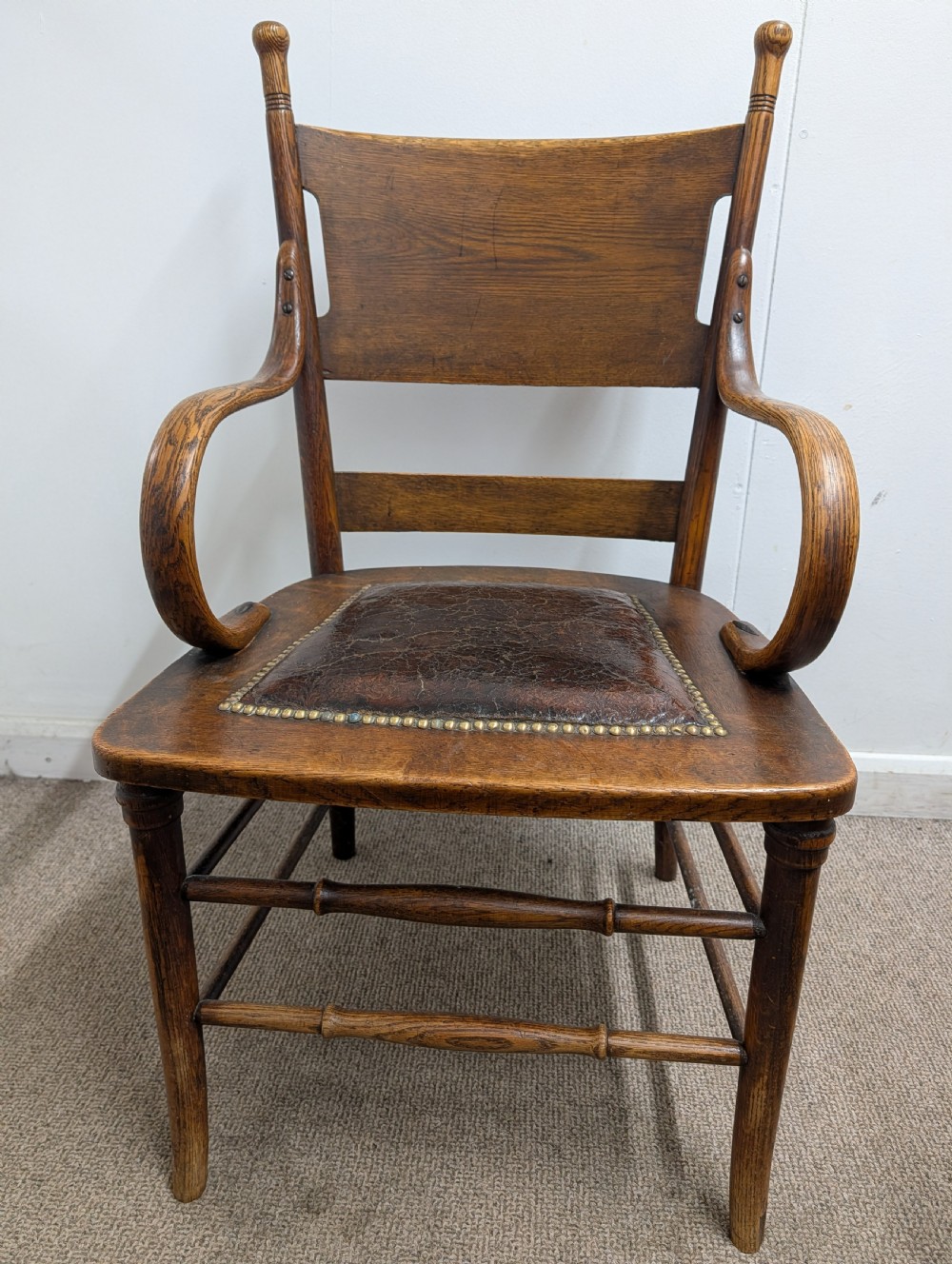 An Arts and Crafts oak open armchair featuring curved sweeping arms, a shaped timber backboard, turned legs with double stretchers, and an inset brown leather seat fixed with brass studs. View XIII