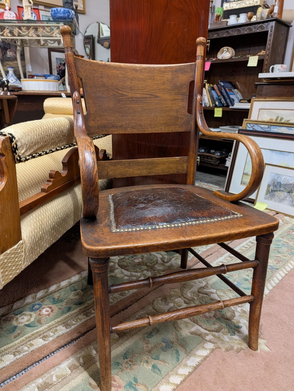 An Arts and Crafts oak open armchair featuring curved sweeping arms, a shaped timber backboard, turned legs with double stretchers, and an inset brown leather seat fixed with brass studs. View XIV