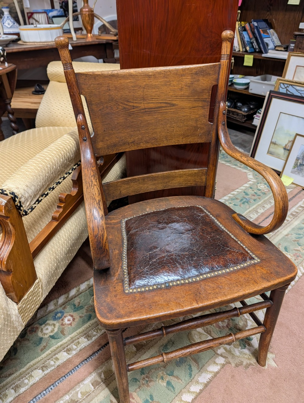 An Arts and Crafts oak open armchair featuring curved sweeping arms, a shaped timber backboard, turned legs with double stretchers, and an inset brown leather seat fixed with brass studs. View XV