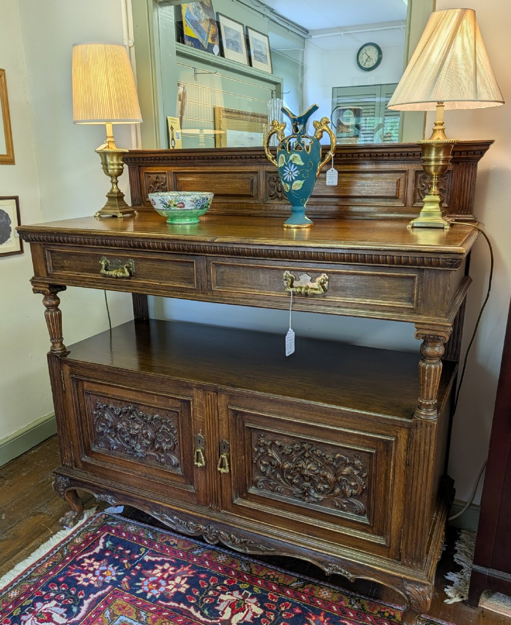 A late Victorian carved oak buffet featuring two top drawers with brass drop handles, an open tier, and a lower double-door cabinet decorated with detailed carved foliage.