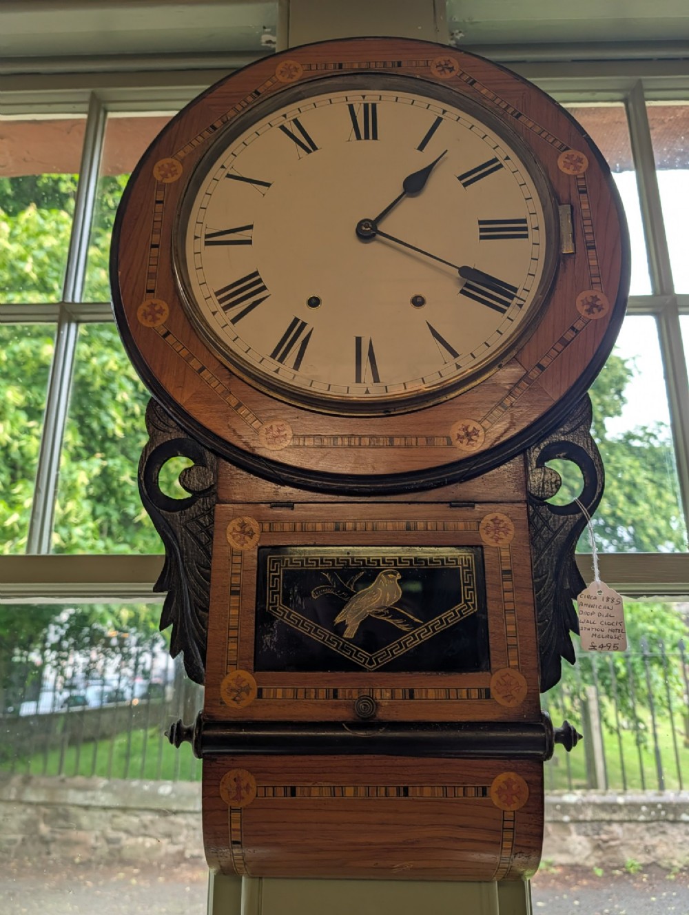 A 19th-century American drop dial wall clock made of inlaid wood with a cream dial, Roman numerals, and a glass-fronted lower panel featuring a bird design.
