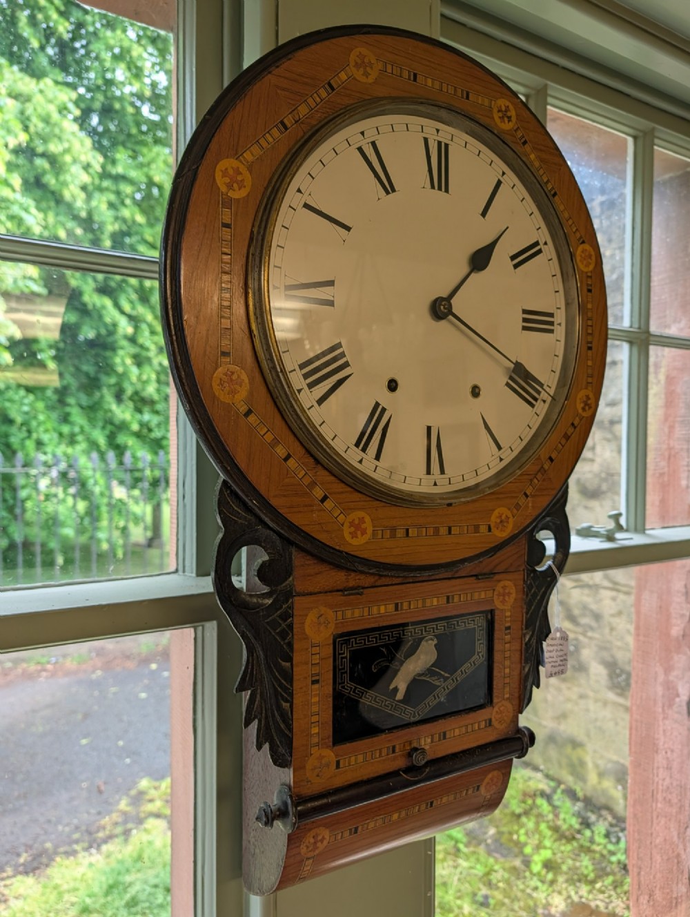 A 19th-century American drop dial wall clock made of inlaid wood with a cream dial, Roman numerals, and a glass-fronted lower panel featuring a bird design. View V