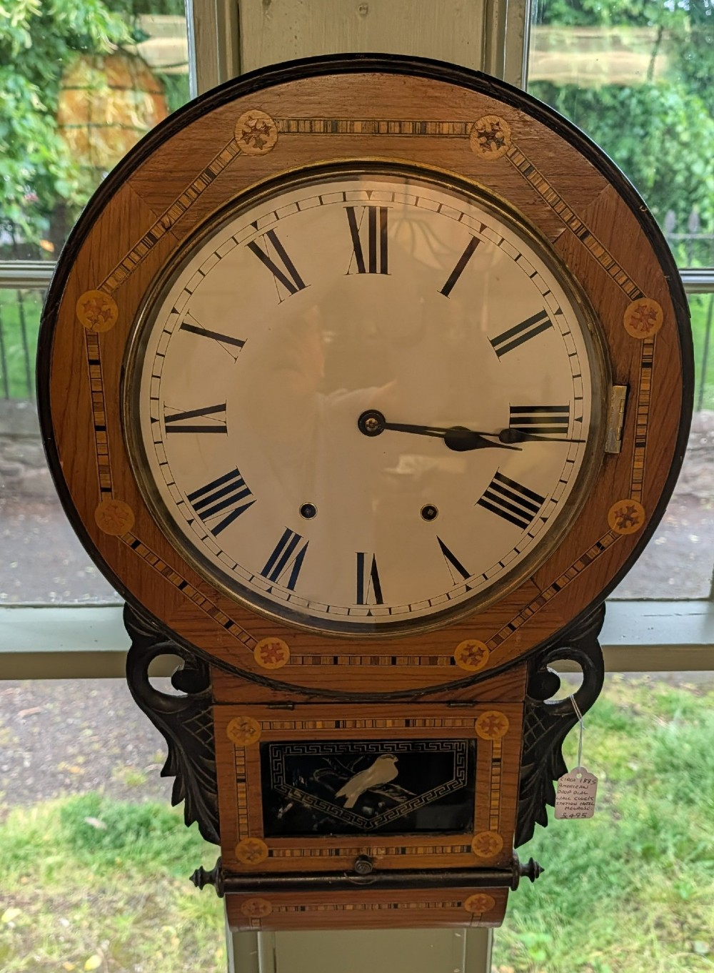 A 19th-century American drop dial wall clock made of inlaid wood with a cream dial, Roman numerals, and a glass-fronted lower panel featuring a bird design. View VI