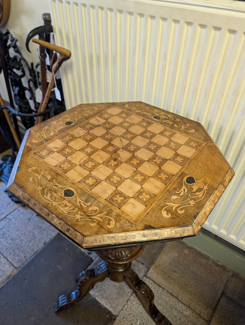 A Victorian octagonal walnut games table featuring an inlaid chessboard lid above a tapering trumpet pedestal on carved tripod legs. View IX