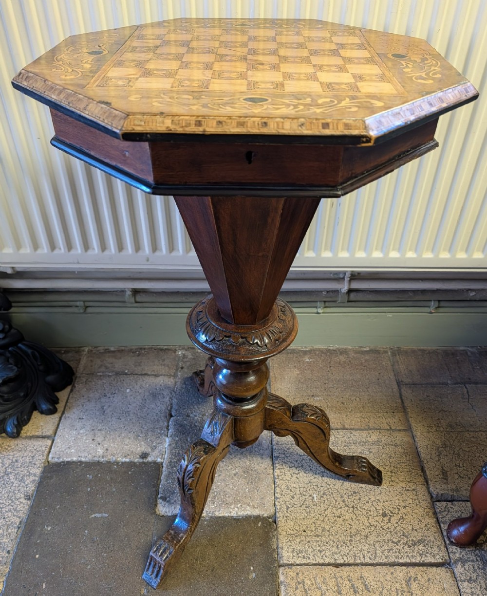 A Victorian octagonal walnut games table featuring an inlaid chessboard lid above a tapering trumpet pedestal on carved tripod legs. View X