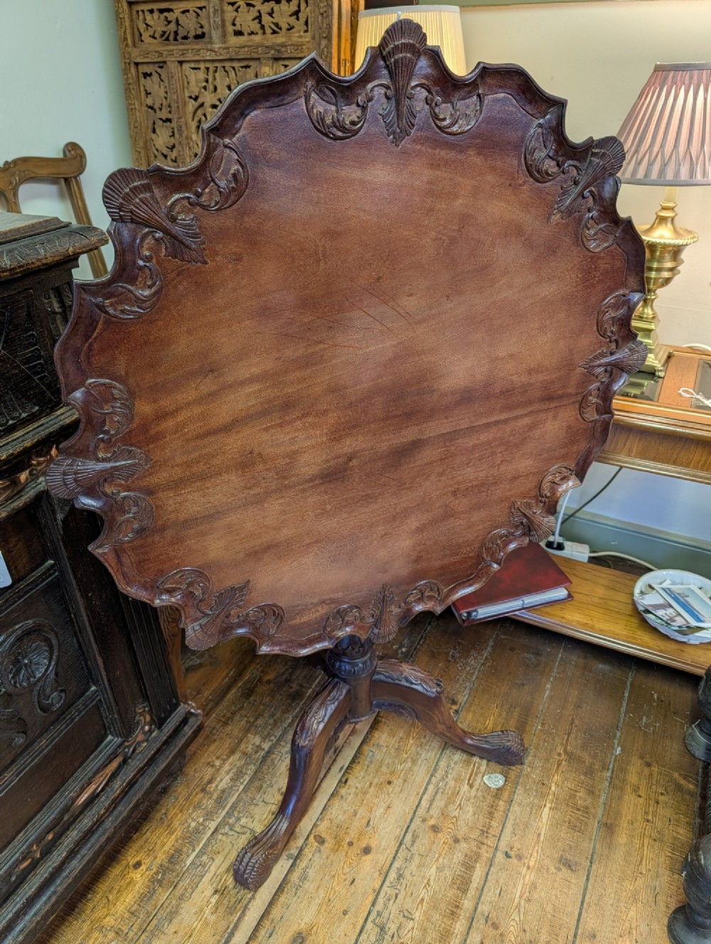 A top-down view of a 19th-century mahogany tilt-top table featuring a carved scalloped edge with shell motifs on a central turned pedestal base. View II