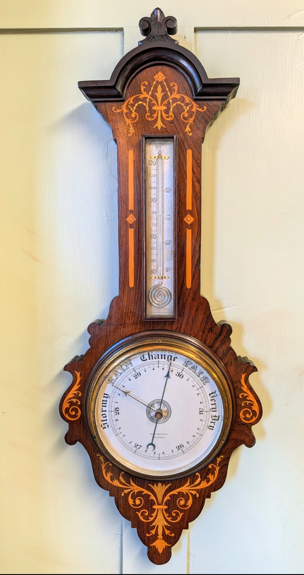 An Edwardian wall-mounted rosewood barometer featuring floral marquetry inlay, a top-mounted glass thermometer, and a circular brass-rimmed pressure dial.