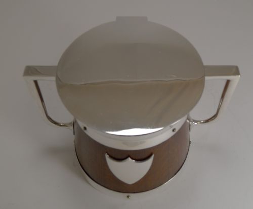 fig 1. A circular English oak biscuit barrel featuring a silver-plated metal rim, flat top cover, central shield plaque, and twin angular side handles.