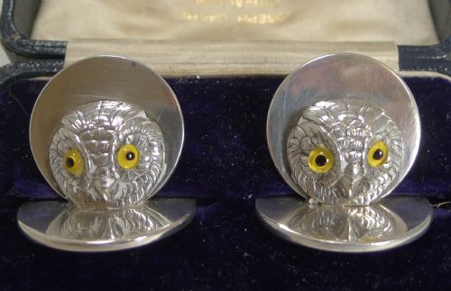 fig 1. A pair of sterling silver menu holders shaped as detailed owl heads with yellow glass eyes resting on circular silver bases inside a blue fitted box.