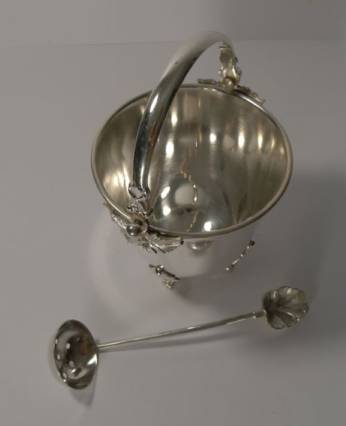 fig 1. A silver-plated ice bucket featuring an overhead swinging handle, leaf-form feet and rim accents, displayed alongside a matching slotted draining spoon.