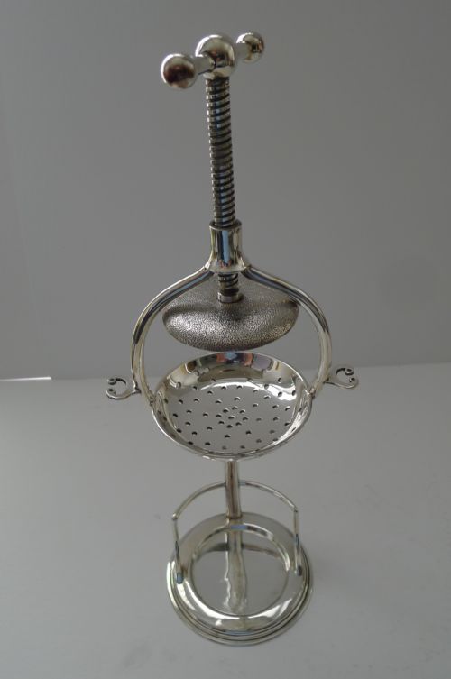 fig 1. A Victorian silver-plated mechanical lemon squeezer featuring a central screw press mechanism positioned over a perforated strainer and clear glass tumbler.