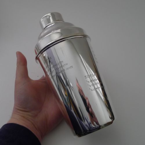 fig 1. A polished silver-plated cocktail shaker featuring engraved classic cocktail recipes on the exterior body.