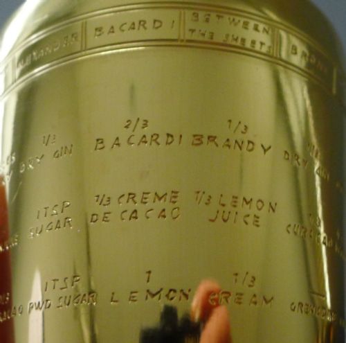 fig 1. A silver-plated Art Deco cocktail shaker featuring a gilded recipe band and rectangular window slots displaying drink ingredients.