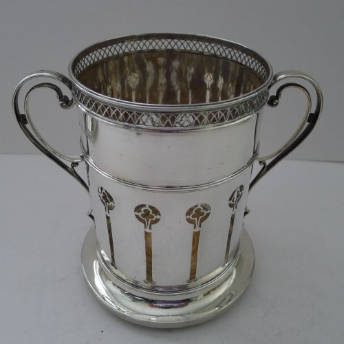 fig 1. An Edwardian sterling silver wine bottle holder featuring pierced vertical motifs, a lattice rim, dual scroll handles, and a circular base.