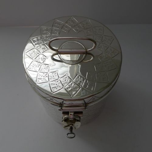 fig 1. A cylindrical silver-plated Victorian biscuit box featuring engraved geometric trellis patterning, a loop top handle, and a hinged clasp with a padlock.