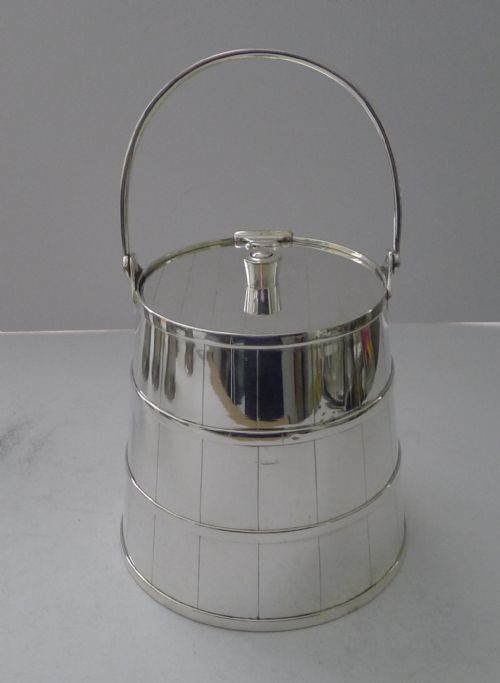 fig 1. A silver-plated cylindrical container styled as a coopered wooden bucket with incised vertical lines, horizontal bands, a flat lid, and a tall curved swing handle.