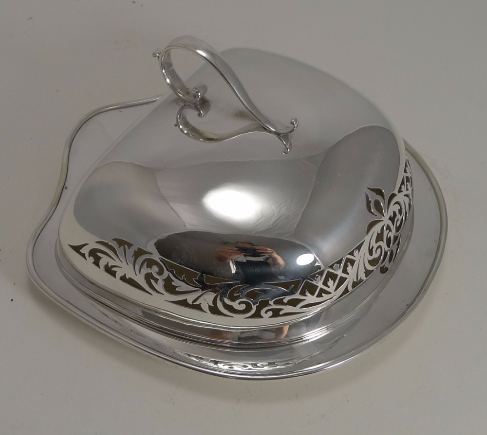 An antique silver-plated cheese dish cover featuring pierced scrollwork decoration, a looped top handle, and a matching fitted tray.