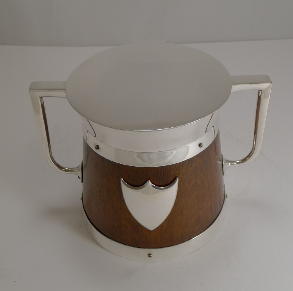 A circular English oak biscuit barrel featuring a silver-plated metal rim, flat top cover, central shield plaque, and twin angular side handles.