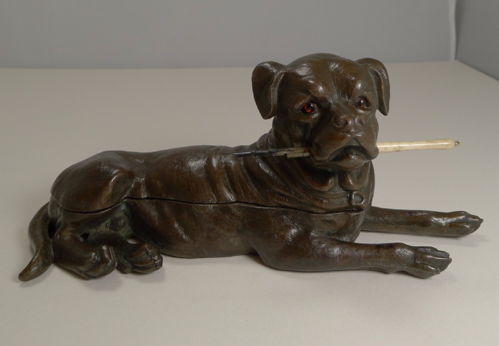 A dark brown patinated bronze inkwell sculpted in the shape of a lying Bullmastiff holding a dip pen in its mouth.