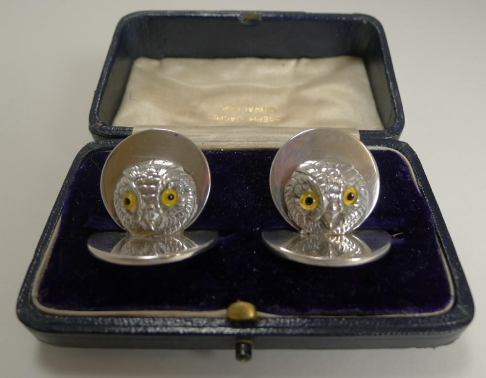 A pair of sterling silver menu holders shaped as detailed owl heads with yellow glass eyes resting on circular silver bases inside a blue fitted box.