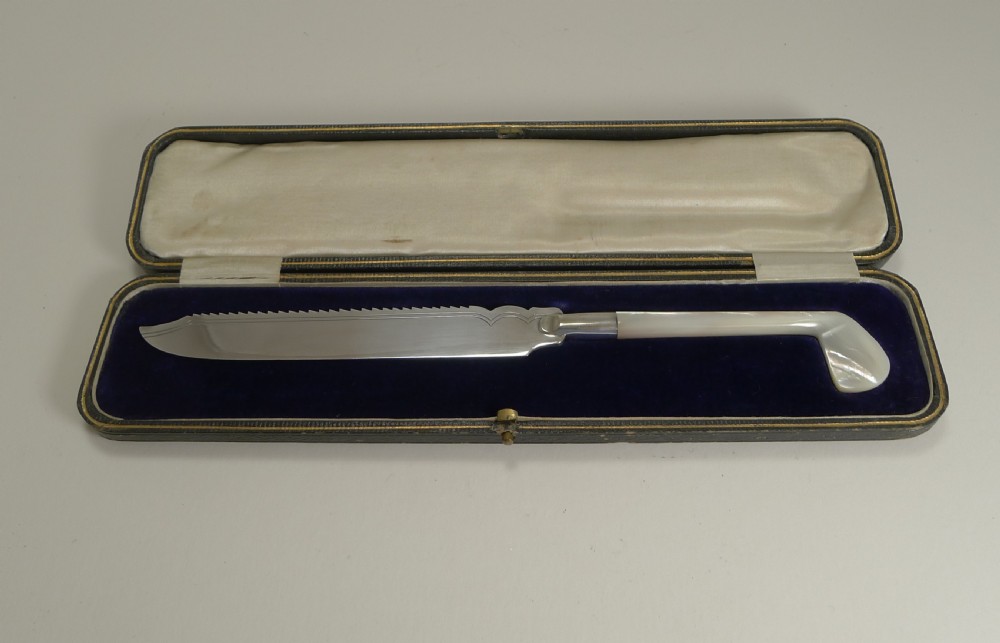 An Edwardian silver-plated cake knife with a serrated edge and a mother of pearl handle shaped like a golf club, displayed inside its fitted velvet presentation case.
