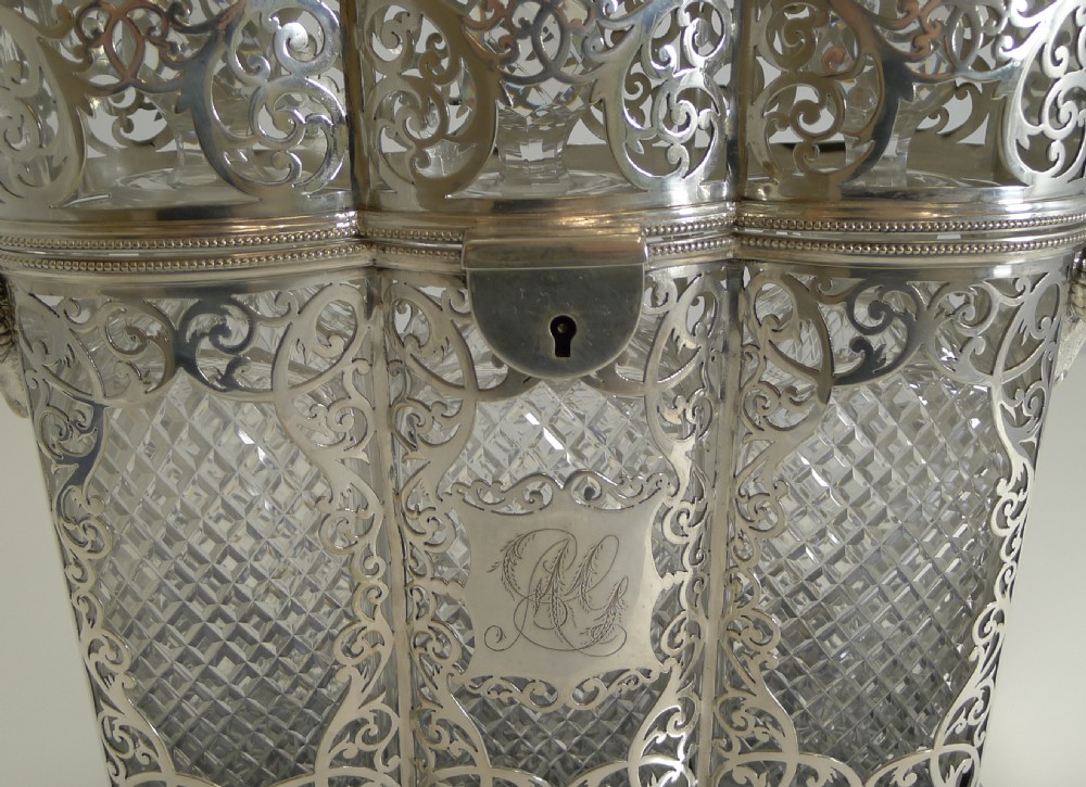 A Victorian silver-plated three-bottle tantalus frame featuring intricate pierced scrollwork, rams head side handles, top carrying handle, and fitted cut crystal decanters. View IV
