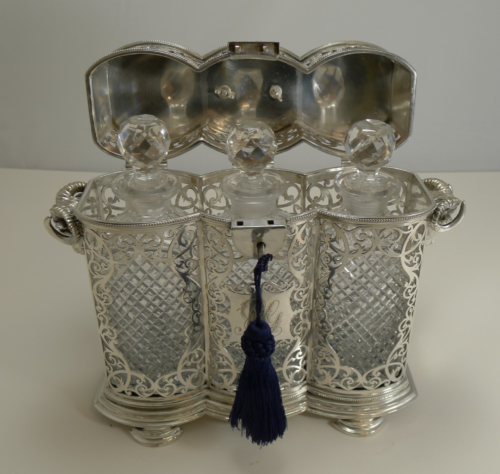 A Victorian silver-plated three-bottle tantalus frame featuring intricate pierced scrollwork, rams head side handles, top carrying handle, and fitted cut crystal decanters. View VIII