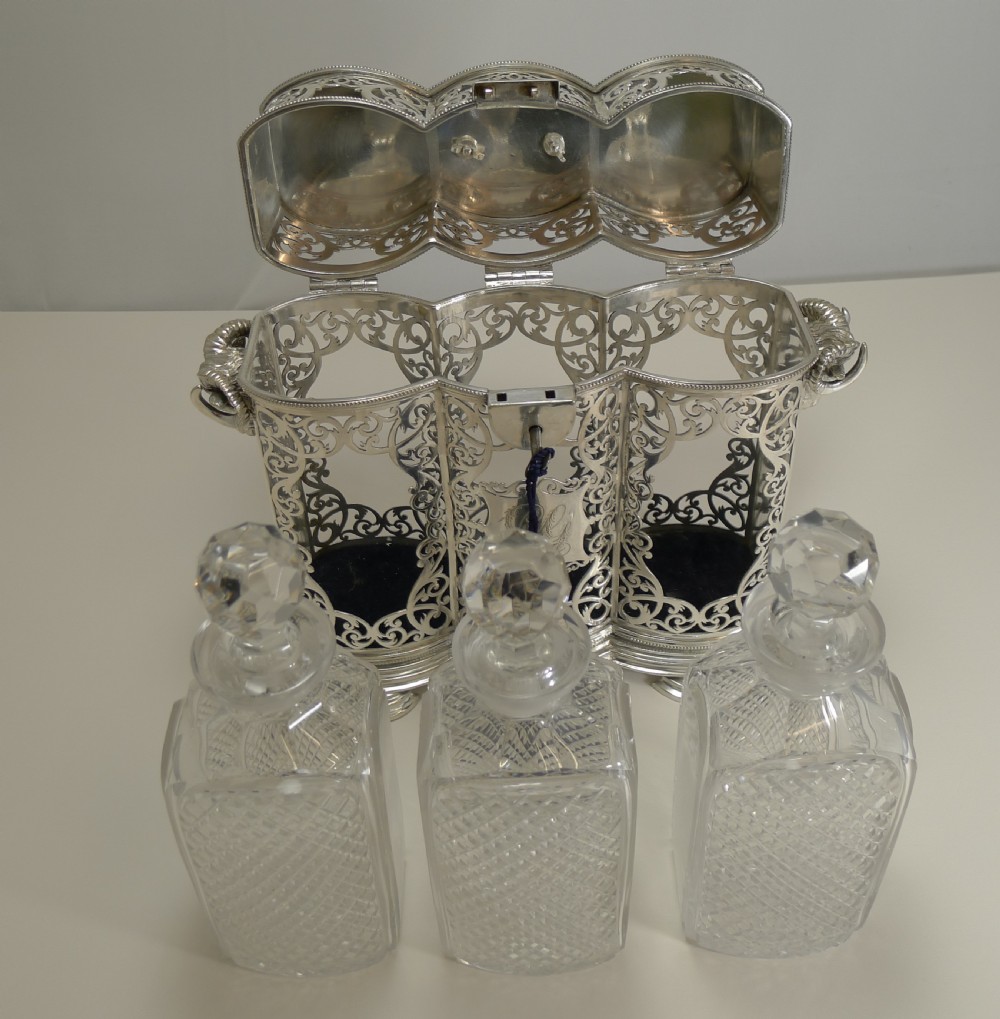 A Victorian silver-plated three-bottle tantalus frame featuring intricate pierced scrollwork, rams head side handles, top carrying handle, and fitted cut crystal decanters. View XI