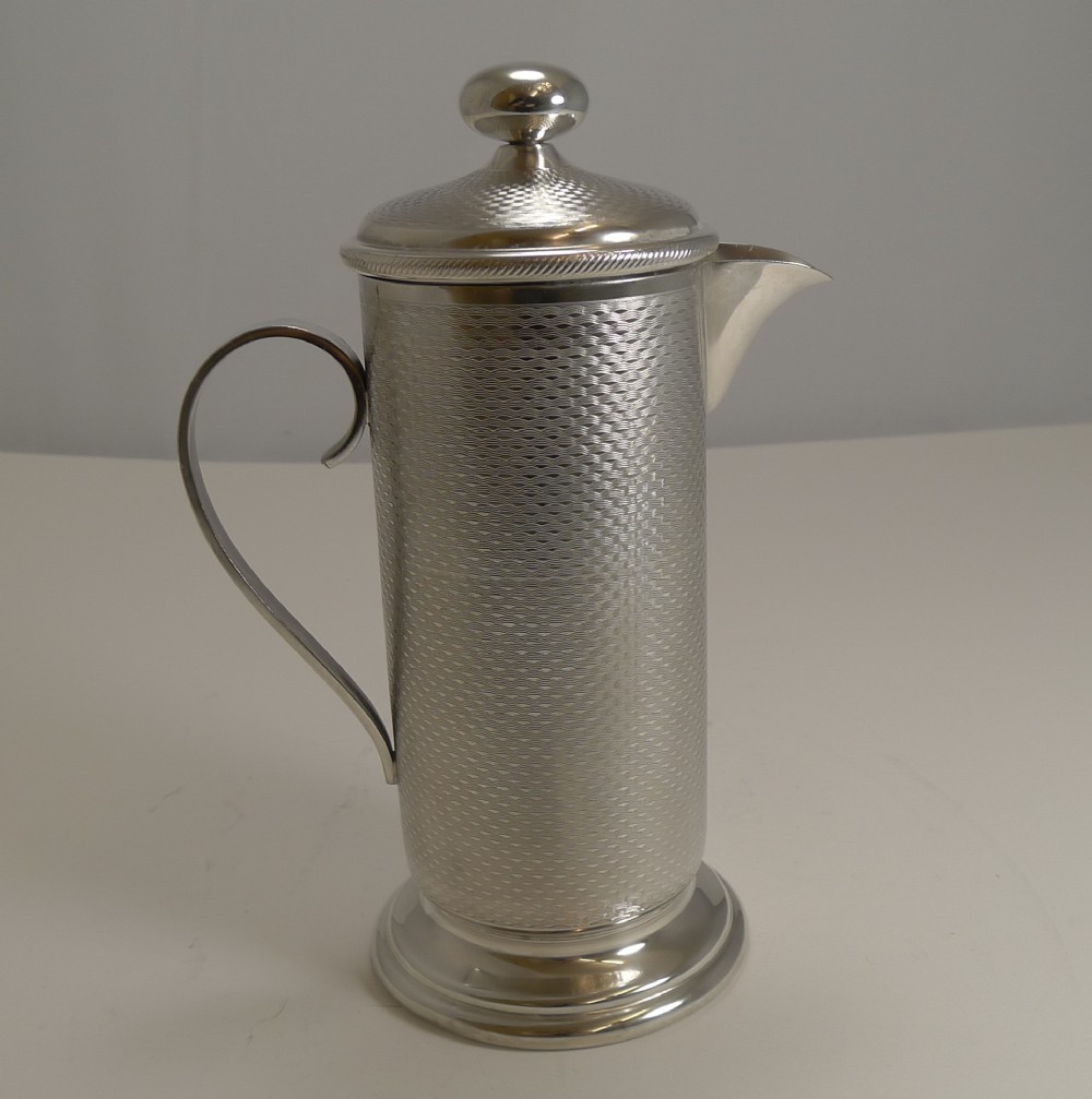 An Art Deco silver-plated cocktail mixer featuring an engine-turned textured body, a curved side handle, a pouring spout, and a lid topped with a spherical finial.