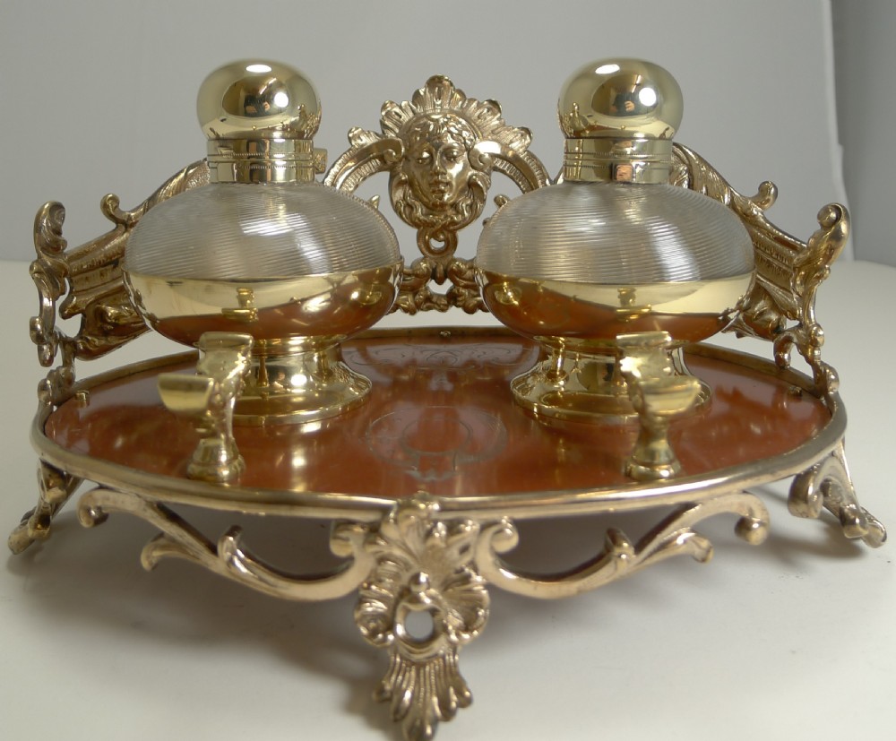An ornate Victorian brass double inkstand featuring two round glass inkwells with brass caps resting on a brass-framed oval base with a classical central mask mount. View IV