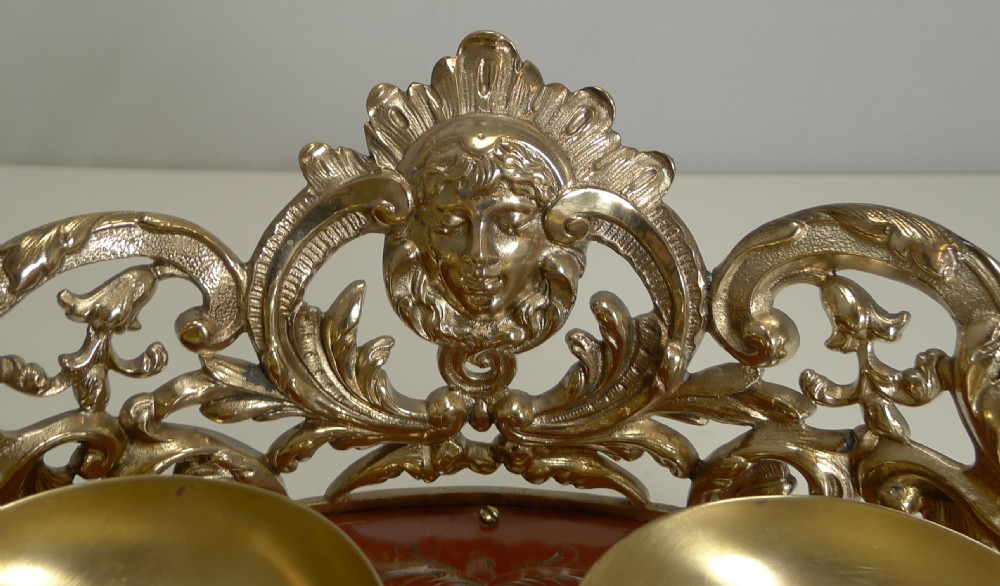 An ornate Victorian brass double inkstand featuring two round glass inkwells with brass caps resting on a brass-framed oval base with a classical central mask mount. View VI