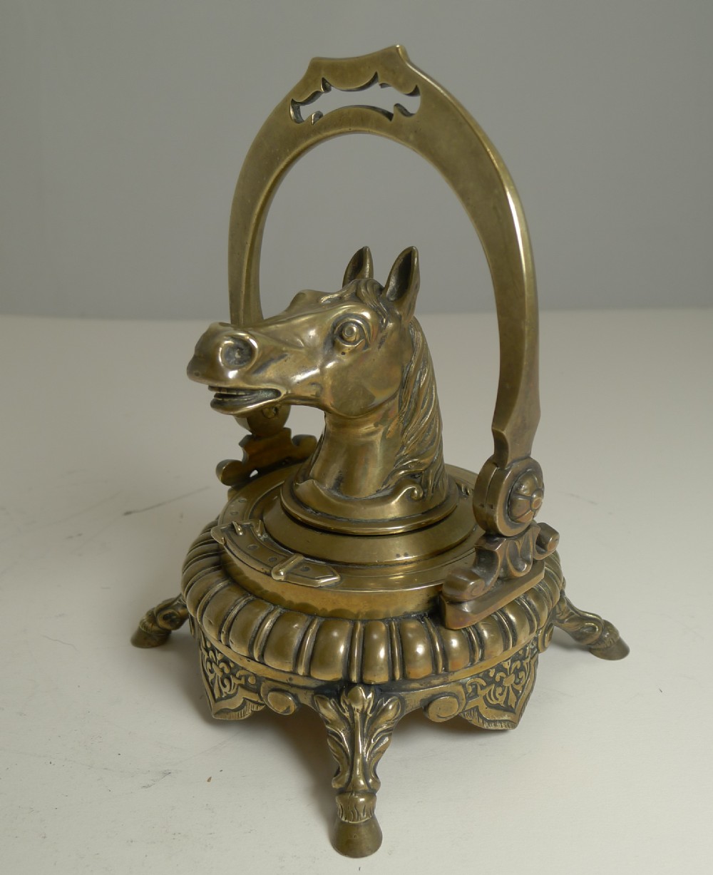 A Victorian cast brass desk inkwell featuring a central sculpted horse head framed by a stirrup arch, resting on a gadrooned base with hoof feet.