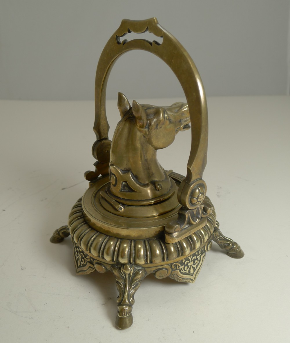 A Victorian cast brass desk inkwell featuring a central sculpted horse head framed by a stirrup arch, resting on a gadrooned base with hoof feet. View V