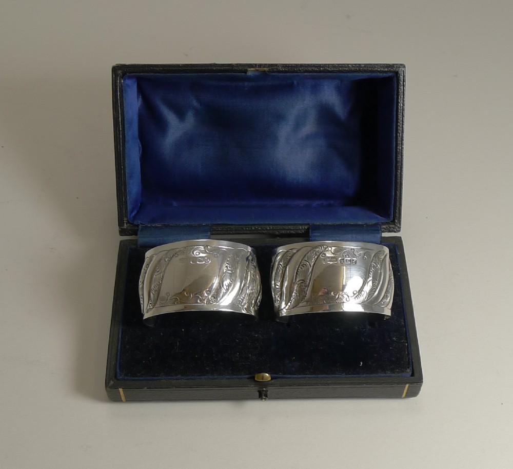 A pair of hallmarked sterling silver napkin rings with scroll engraving, presented inside a fitted black box lined with blue velvet.