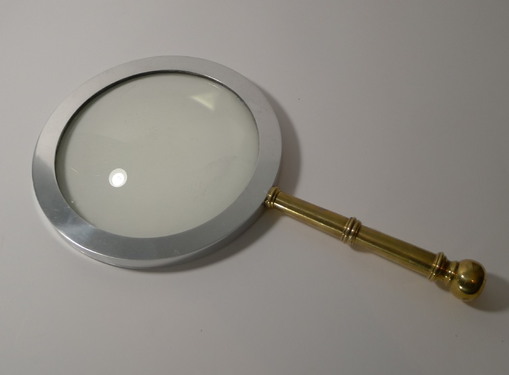 A large round magnifying glass featuring a wide circular silver-toned metal frame around a glass lens and a turned brass handle with a spherical finial. View II
