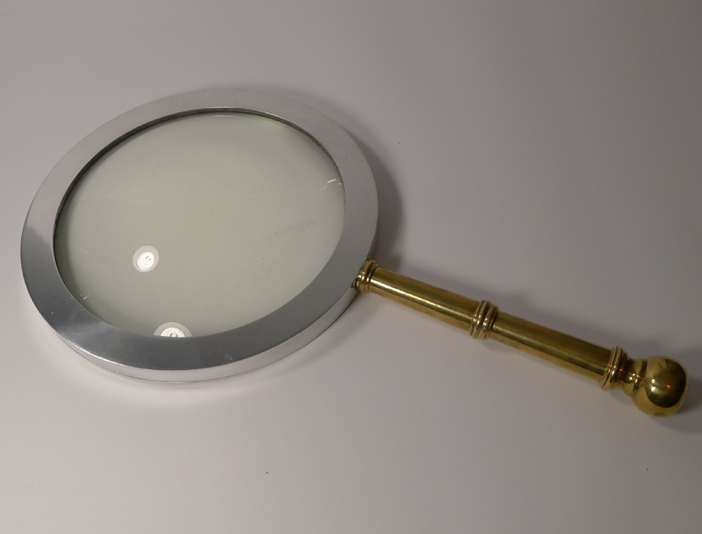 A large round magnifying glass featuring a wide circular silver-toned metal frame around a glass lens and a turned brass handle with a spherical finial. View III