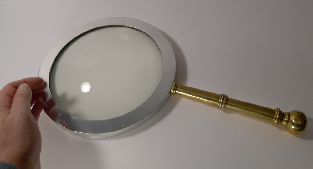 A large round magnifying glass featuring a wide circular silver-toned metal frame around a glass lens and a turned brass handle with a spherical finial. View IV