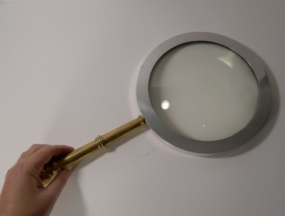A large round magnifying glass featuring a wide circular silver-toned metal frame around a glass lens and a turned brass handle with a spherical finial. View V